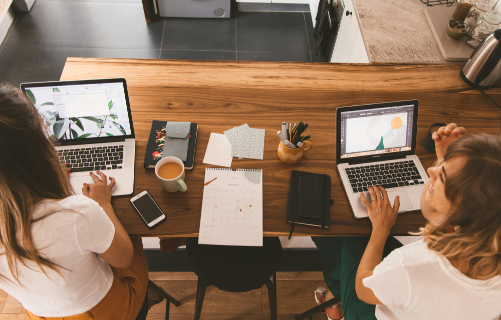 An overhead shot of two women working on their laptop with their coffee, notebook and pens on their table