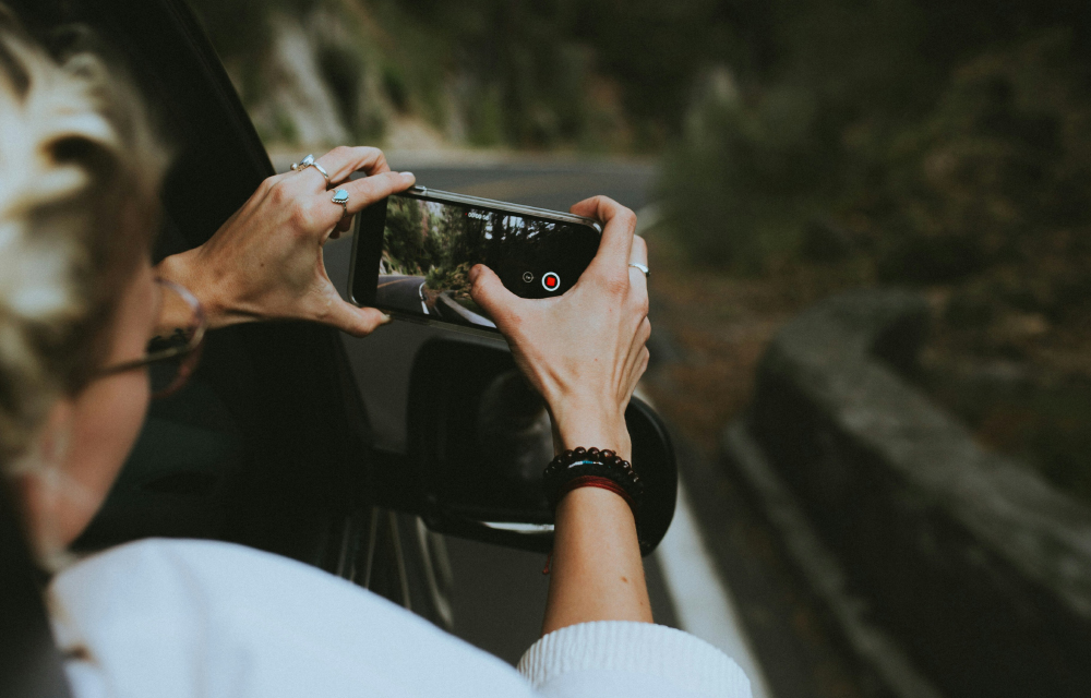 A woman inside a car, holding a phone in her hand, reached out the car window to capture or record the road in front of them.