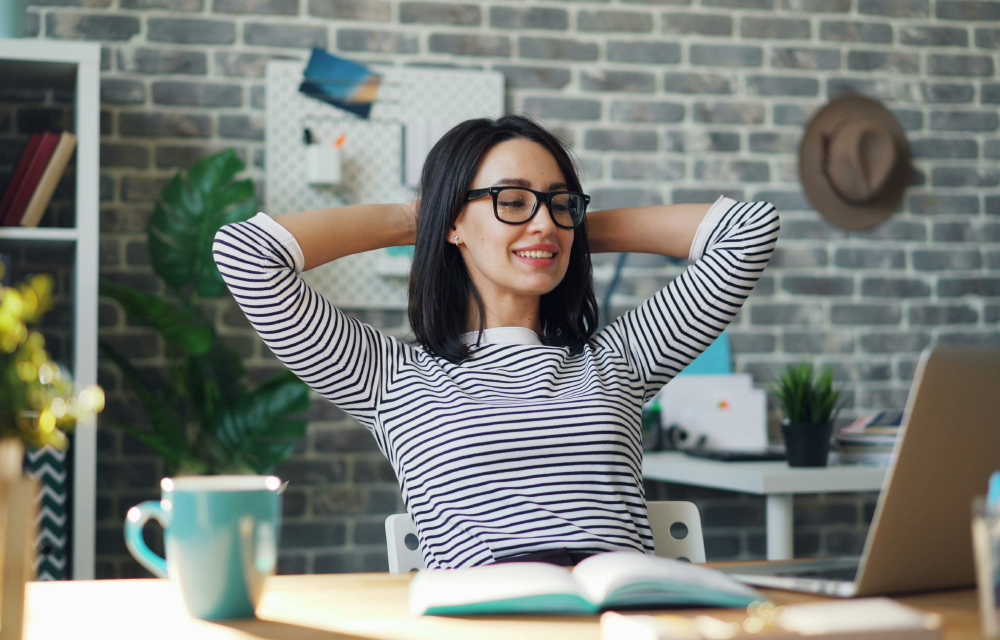 A woman with glasses is seated at a desk, her arms raised, expressing enthusiasm and relief after finishing a task.