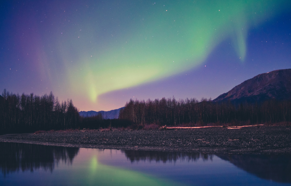 Northern lights illuminate the night sky over a mountain and forest, reflecting in a calm river below.