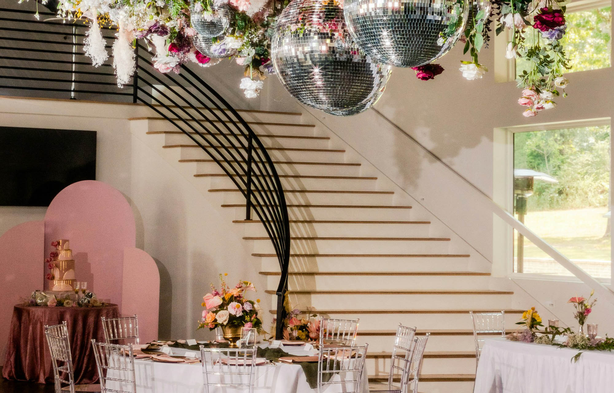 Elegant event venue with a sweeping staircase and black railing, white dining tables, floral centerpieces, and mirrored disco balls for a festive mood.